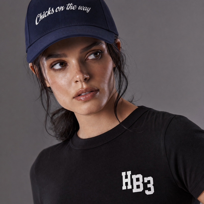 A woman wearing a black short-sleeve t-shirt with a white 'HB3' logo on the chest and a navy blue baseball cap with white cursive text, photographed against a solid gray studio background.